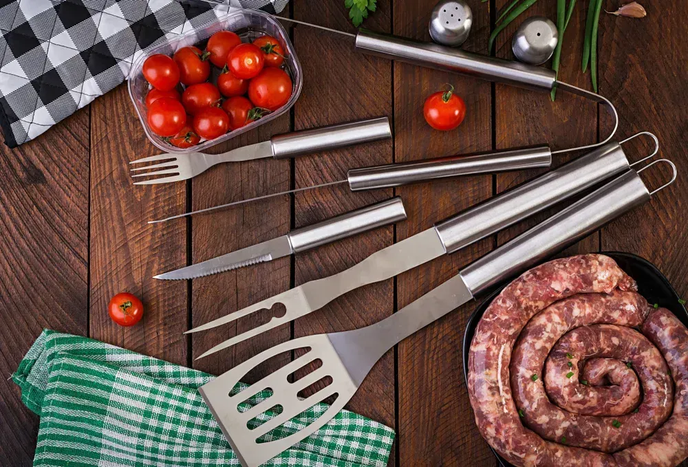 Barbecue tools and ingredients on a wooden surface: sausage, tomatoes, and herbs. — Pigglys Supermarket, Takeaway & Bottle Shop in The Gap, NT