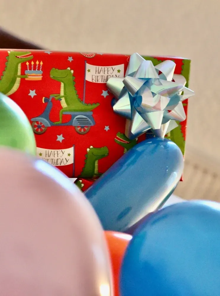 Birthday gift wrapped in dinosaur paper with a blue bow, surrounded by colourful balloons. — Pigglys Supermarket, Takeaway & Bottle Shop in The Gap, NT