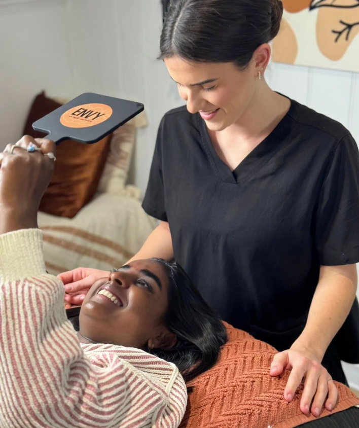 a woman client looking at herself in the mirror and smiling while a beauty therapist smiles looking down at her
 — Envy Cosmetic Collective in Maitland, NSW