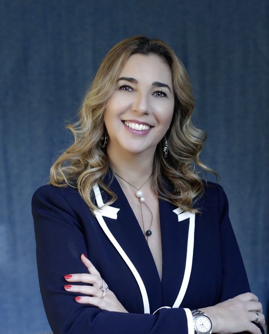 A woman in a blue jacket and white trim is smiling with her arms crossed.