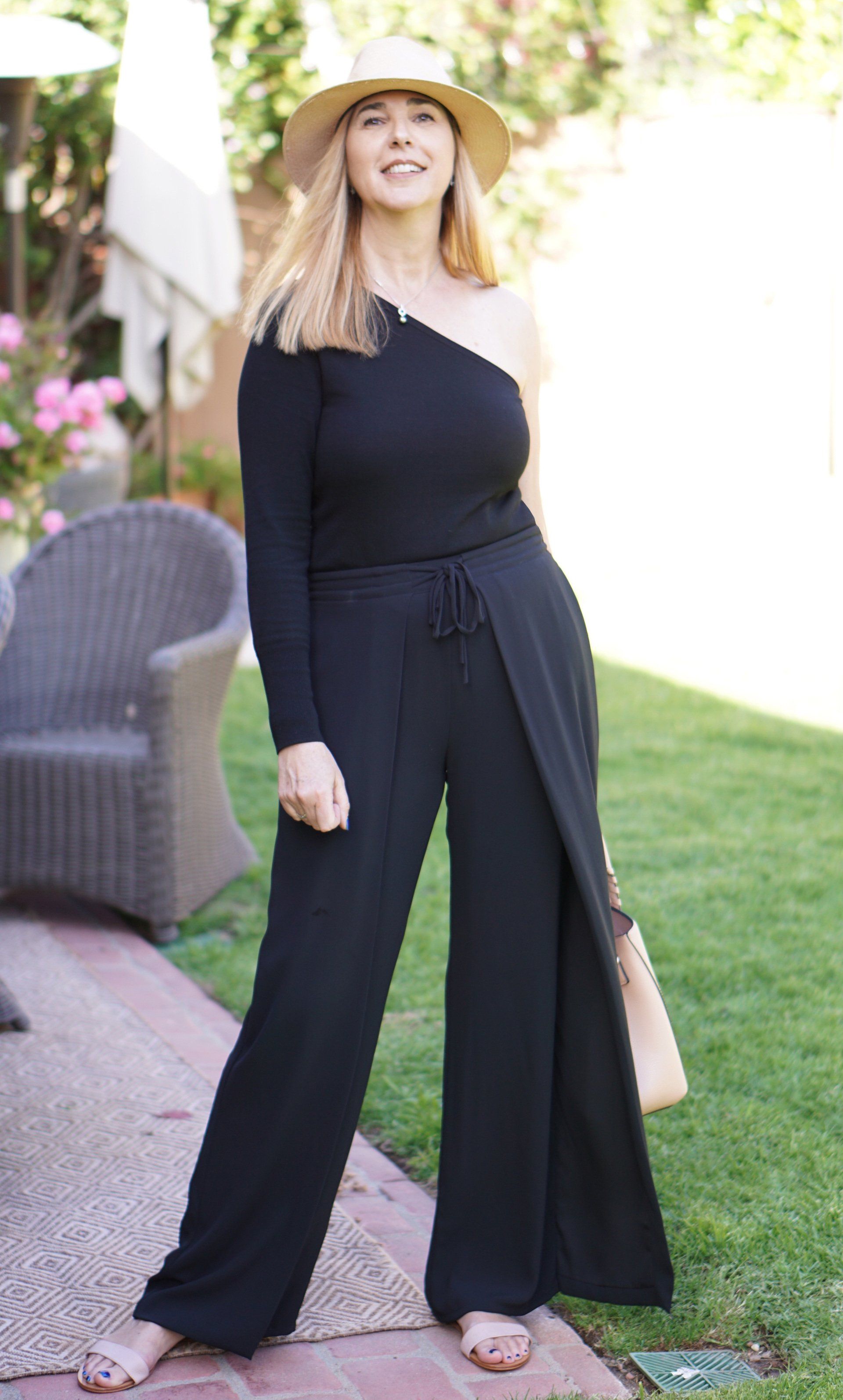 A woman wearing a black jumpsuit and a hat is standing in a yard.