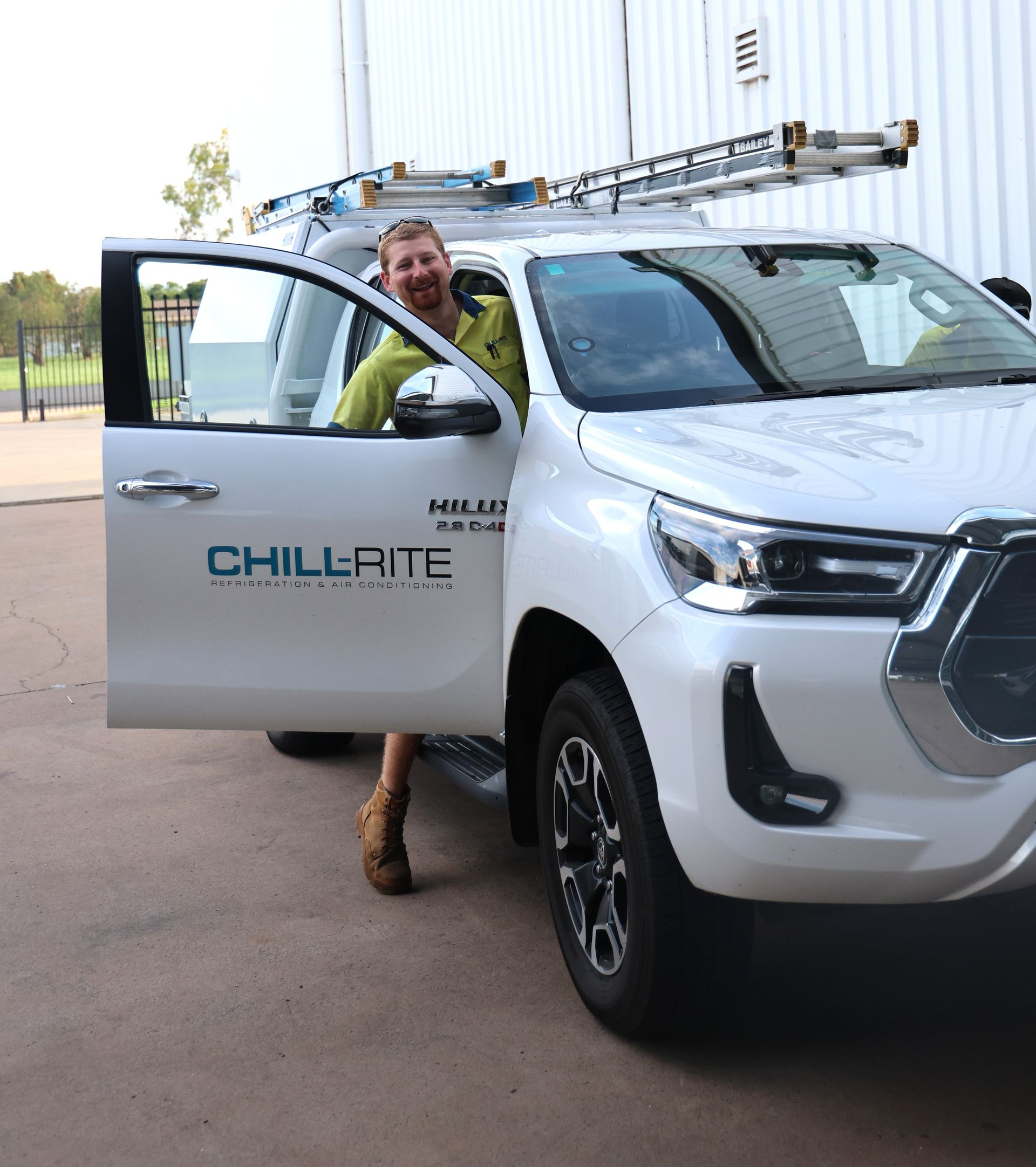 Man smiling beside white company truck, door open. Ladder on roof, building in background -— Chill-Rite Refrigeration and Air Conditioning in Dubbo, NSW