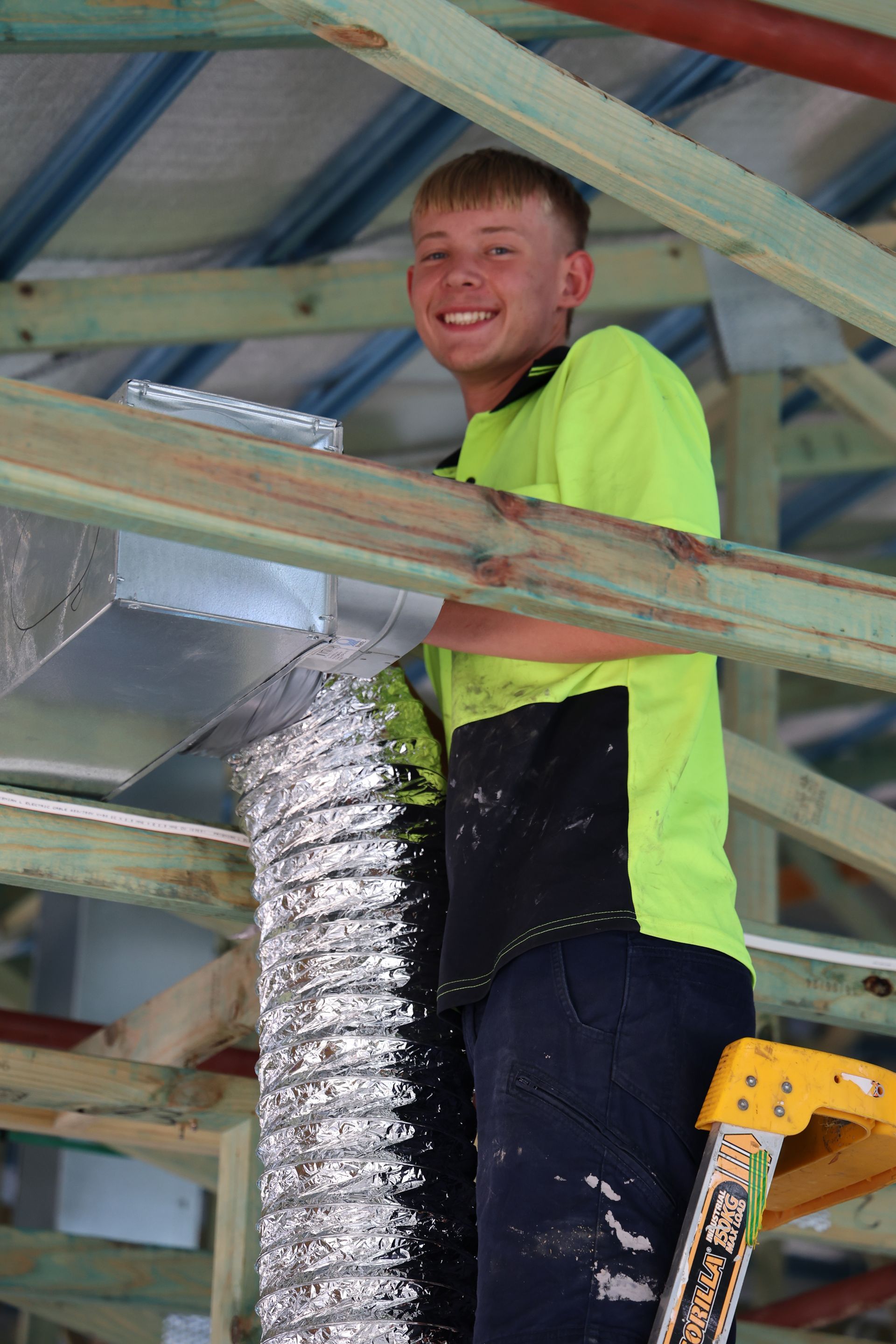 Smiling person in a construction setting, connecting ductwork. Wearing safety vest, standing on a ladder— Chill-Rite Refrigeration and Air Conditioning in Newcastle, NSW