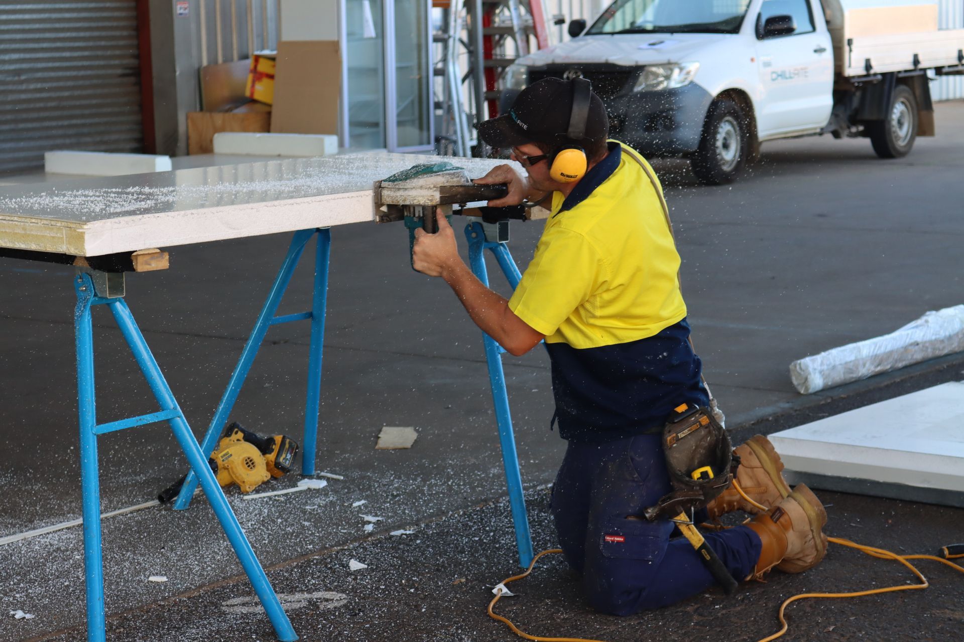 Construction worker kneeling, sawing building material on sawhorses, wearing ear protection, yellow shirt, and tool belt— Chill-Rite Refrigeration and Air Conditioning in Dubbo, NSW