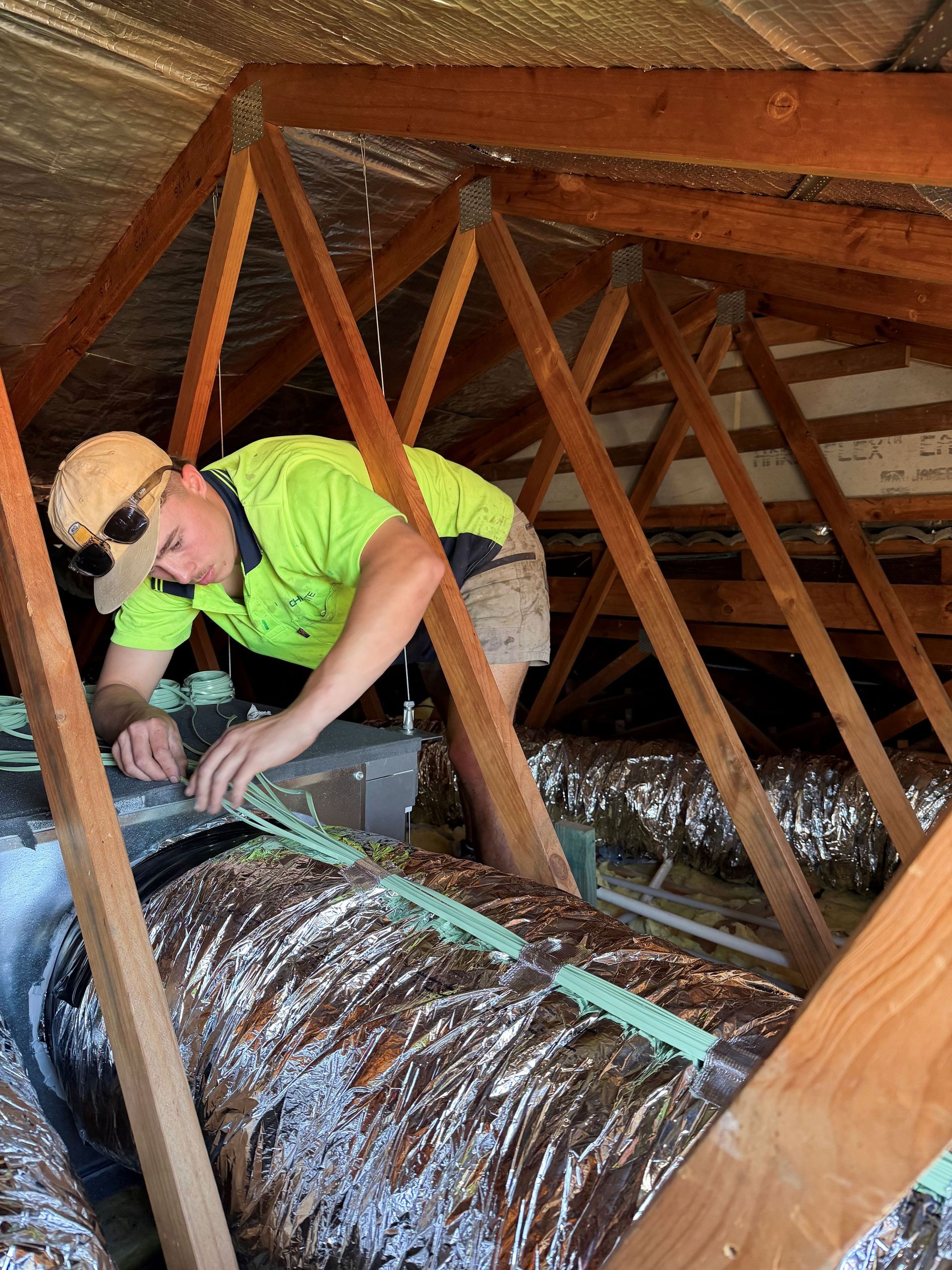 Person in a bright vest working on ductwork inside an attic, surrounded by wooden beams and insulation— Chill-Rite Refrigeration and Air Conditioning in Dubbo, NSW
