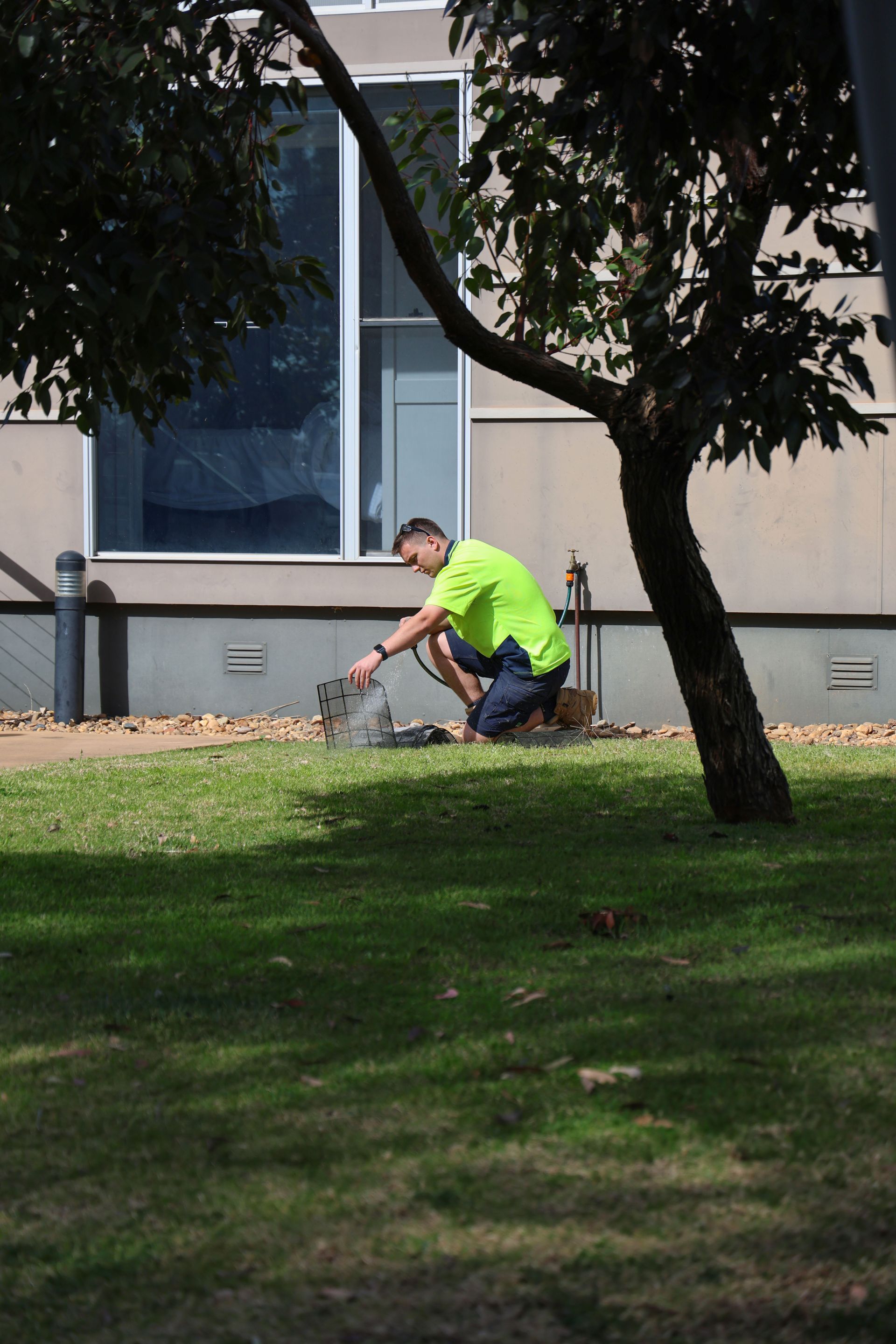 Man in a neon shirt kneeling on grass by a tree, working near a building's wall— Chill-Rite Refrigeration and Air Conditioning in Dubbo, NSW