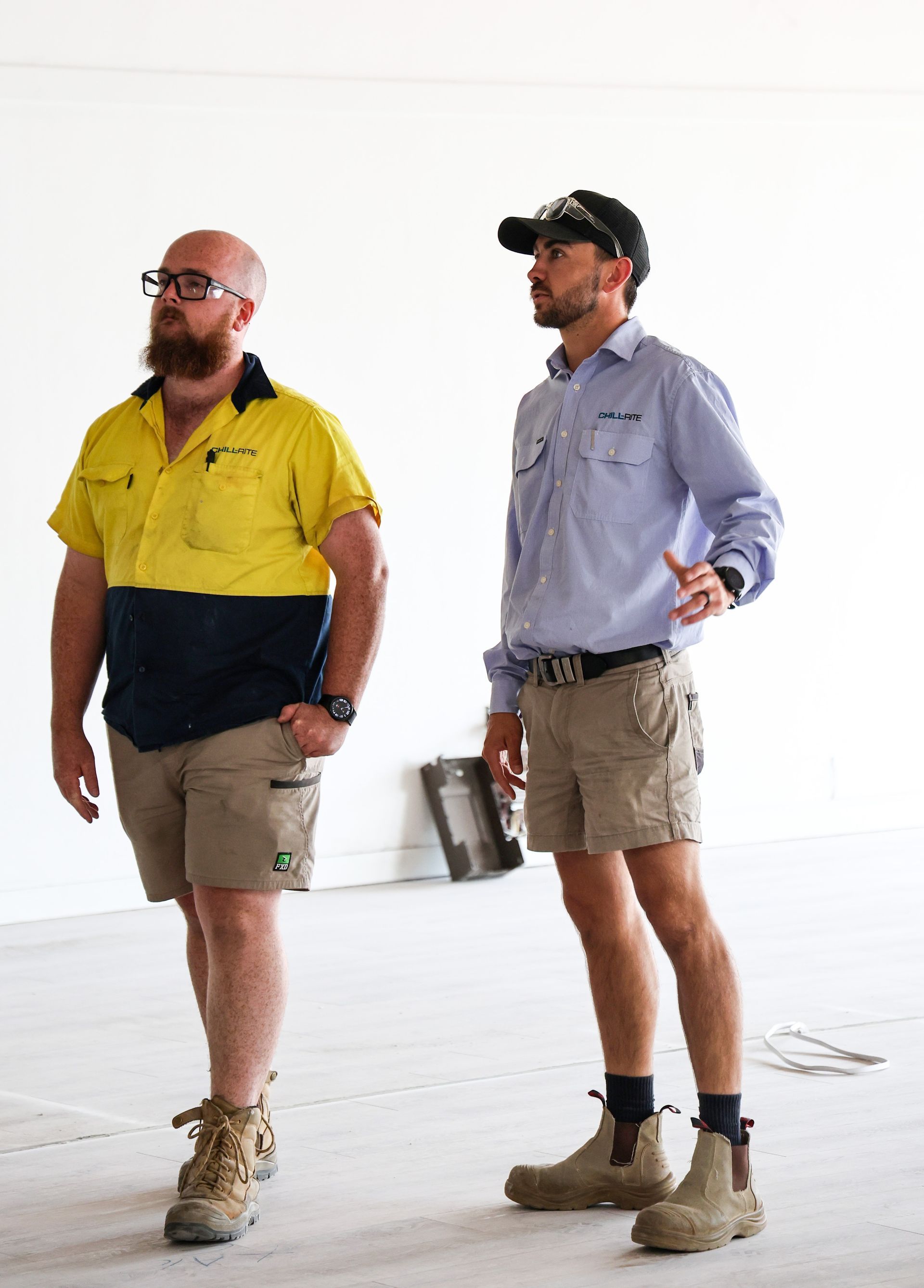 Two men in work clothes standing in an unfinished building. One gestures while the other looks on— Chill-Rite Refrigeration and Air Conditioning in Dubbo, NSW
