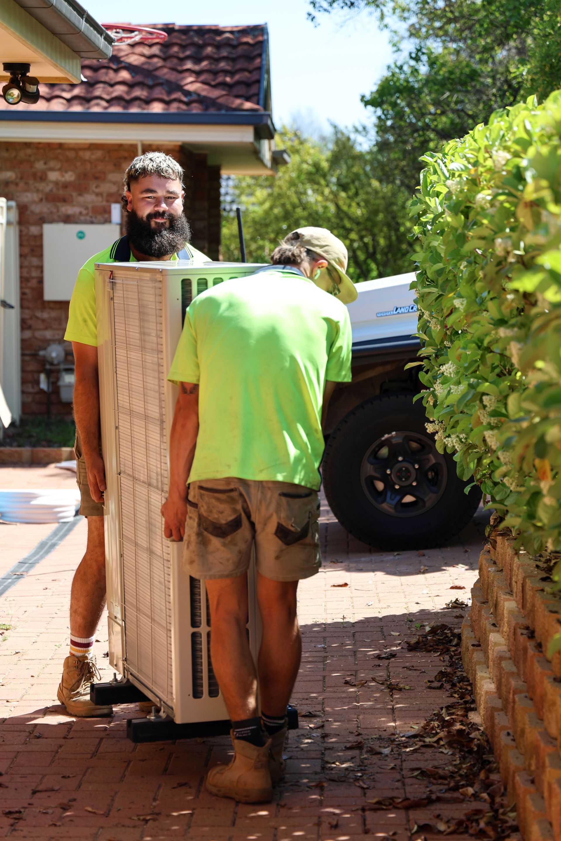 Two workers carrying a large, light-colored rectangular object. One has a beard, all wear work attire. Outdoors next to a brick wall— Chill-Rite Refrigeration and Air Conditioning in Dubbo, NSW