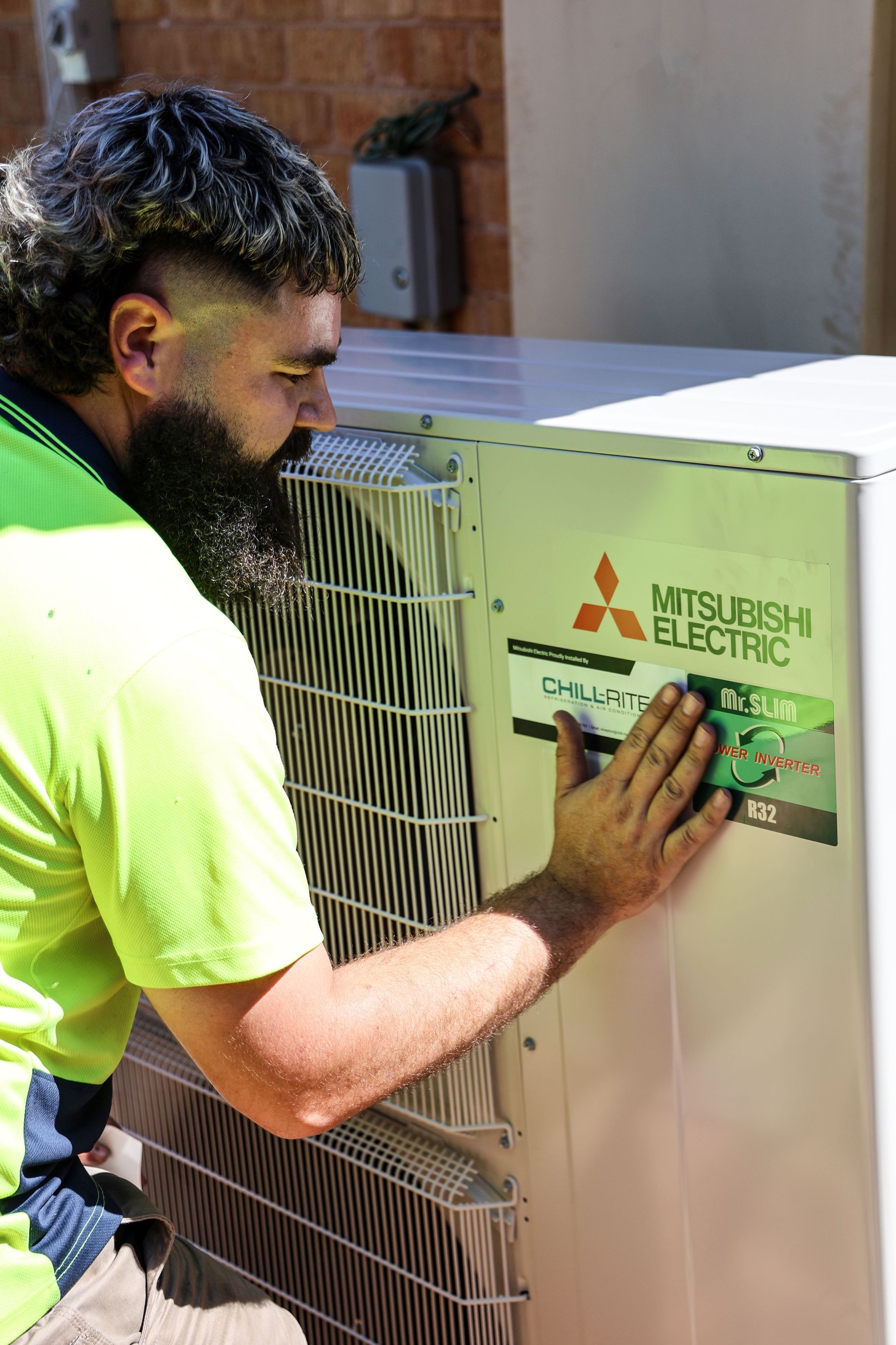 A man with a mullet and beard examines a Mitsubishi air conditioning unit outside.