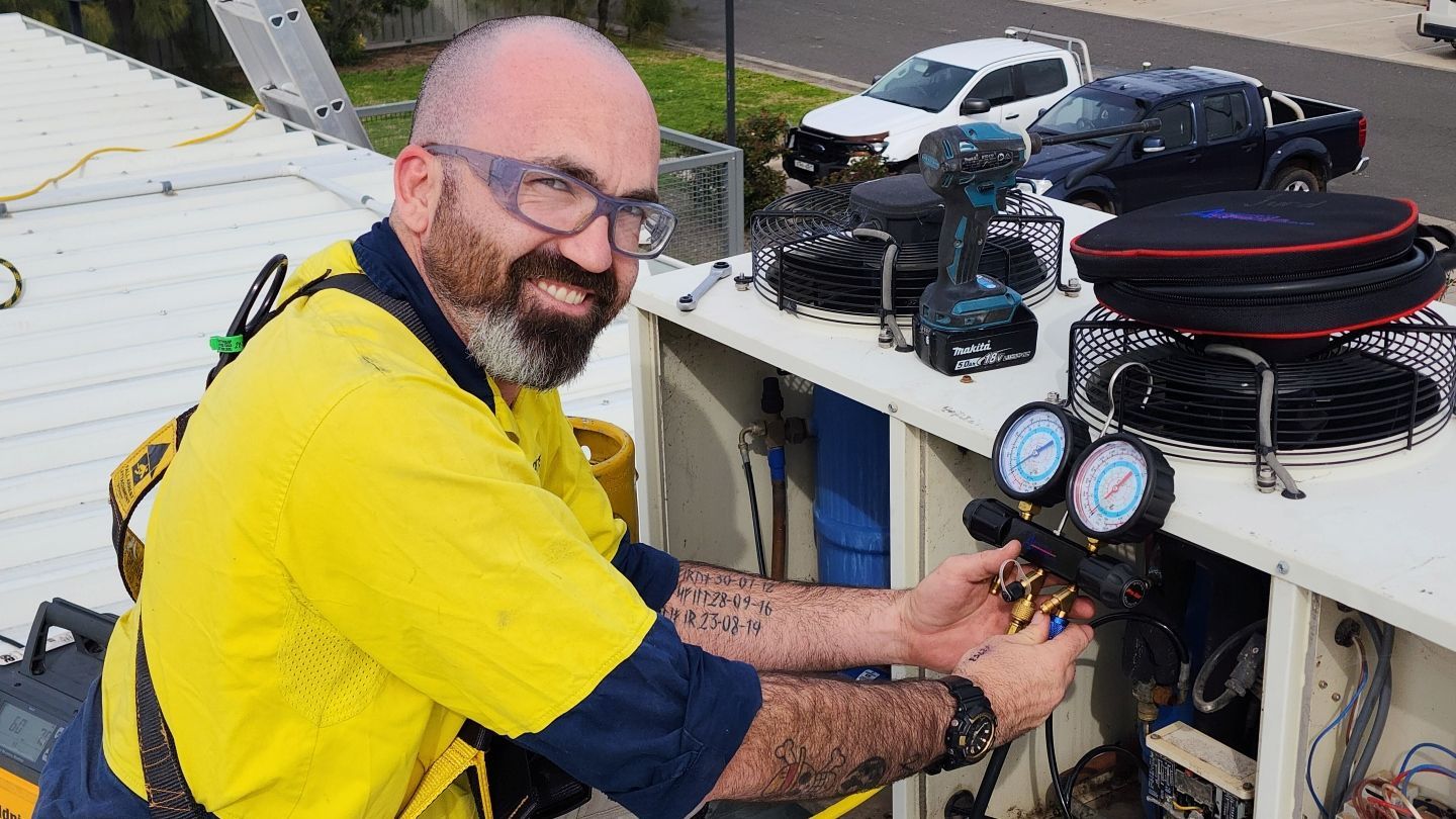 Man on rooftop in safety gear, working on equipment. Smiling, with tools nearby and cars in the background— Chill-Rite Refrigeration and Air Conditioning in Dubbo, NSW