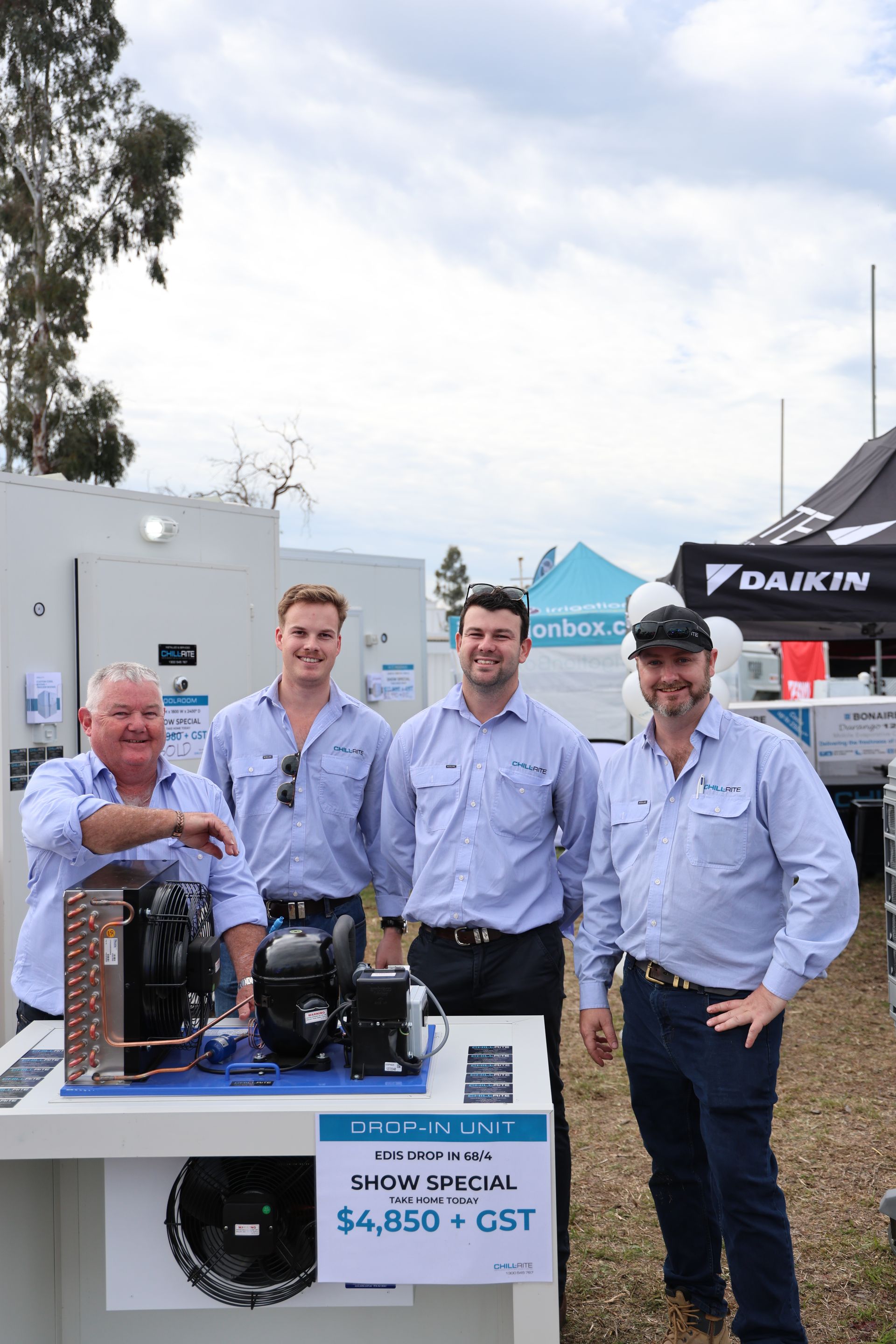 Four men posing by HVAC equipment display at an outdoor event, product sign reading 