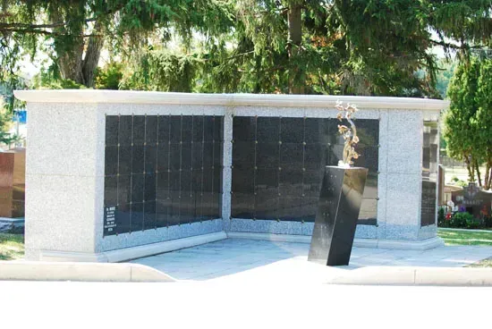 Outdoor memorial wall with dark stone panels and a black sculpture in front, surrounded by trees.