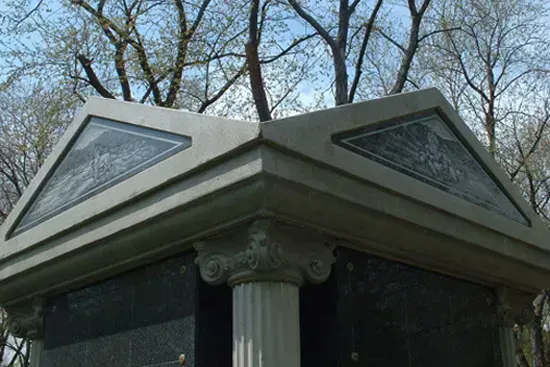Stone mausoleum roof with triangular glass panels and a decorative column against bare trees