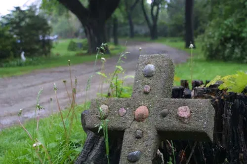 Weathered stone cross in a grassy cemetery, with a blurred tree-lined path in the background.