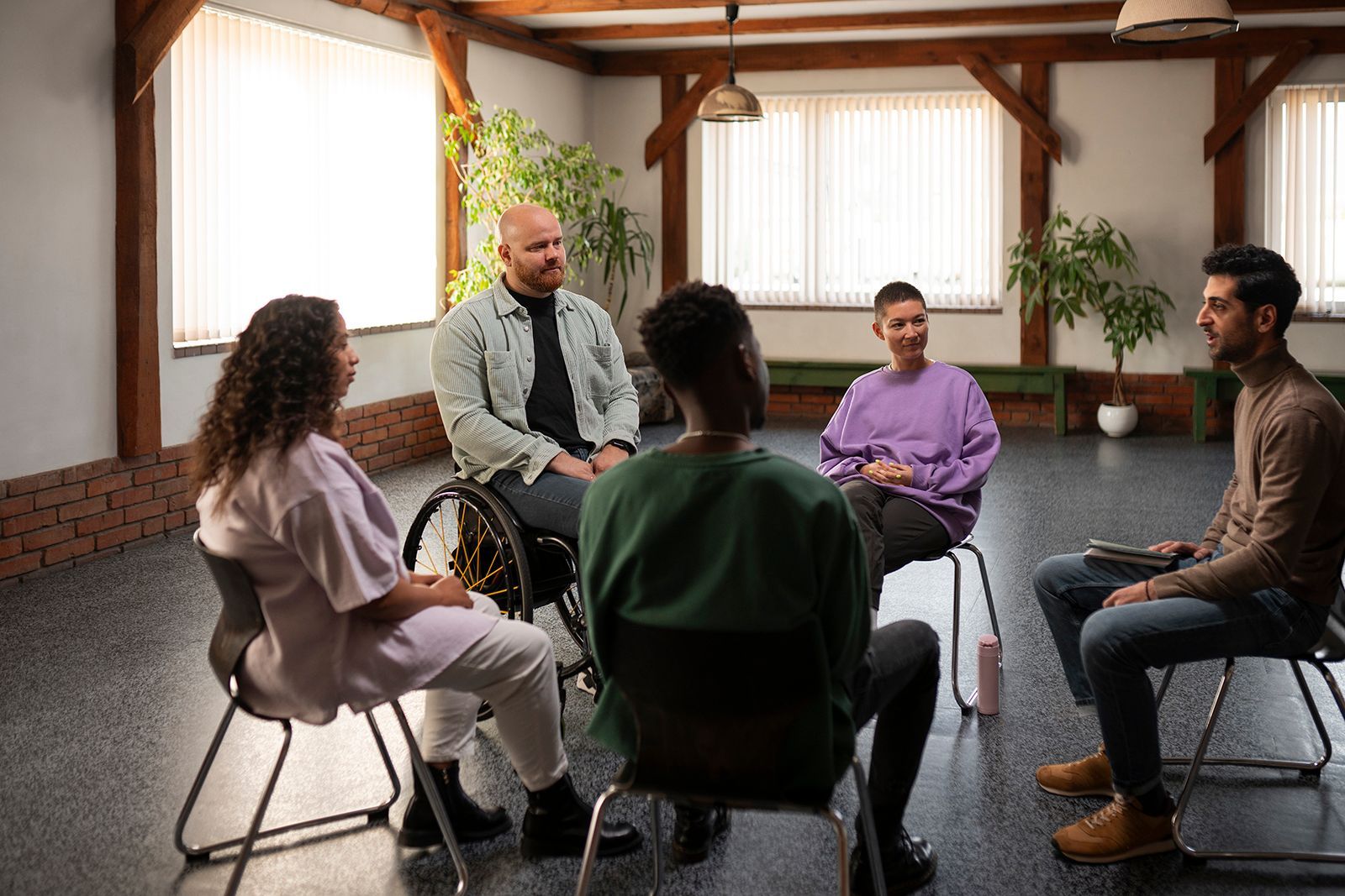 Sesión de terapia grupal en círculo. Personas conversando, expresiones relajadas, en una sala con vigas de madera y plantas.img