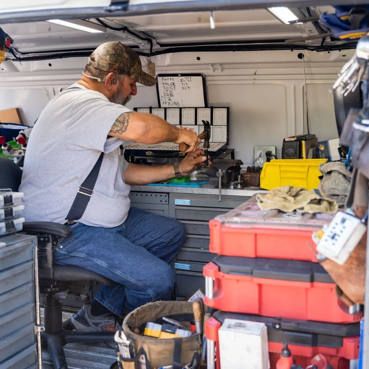 A worker sits at a workbench inside a vehicle, using a hammer to work on a small mechanical part near red tool boxes.
