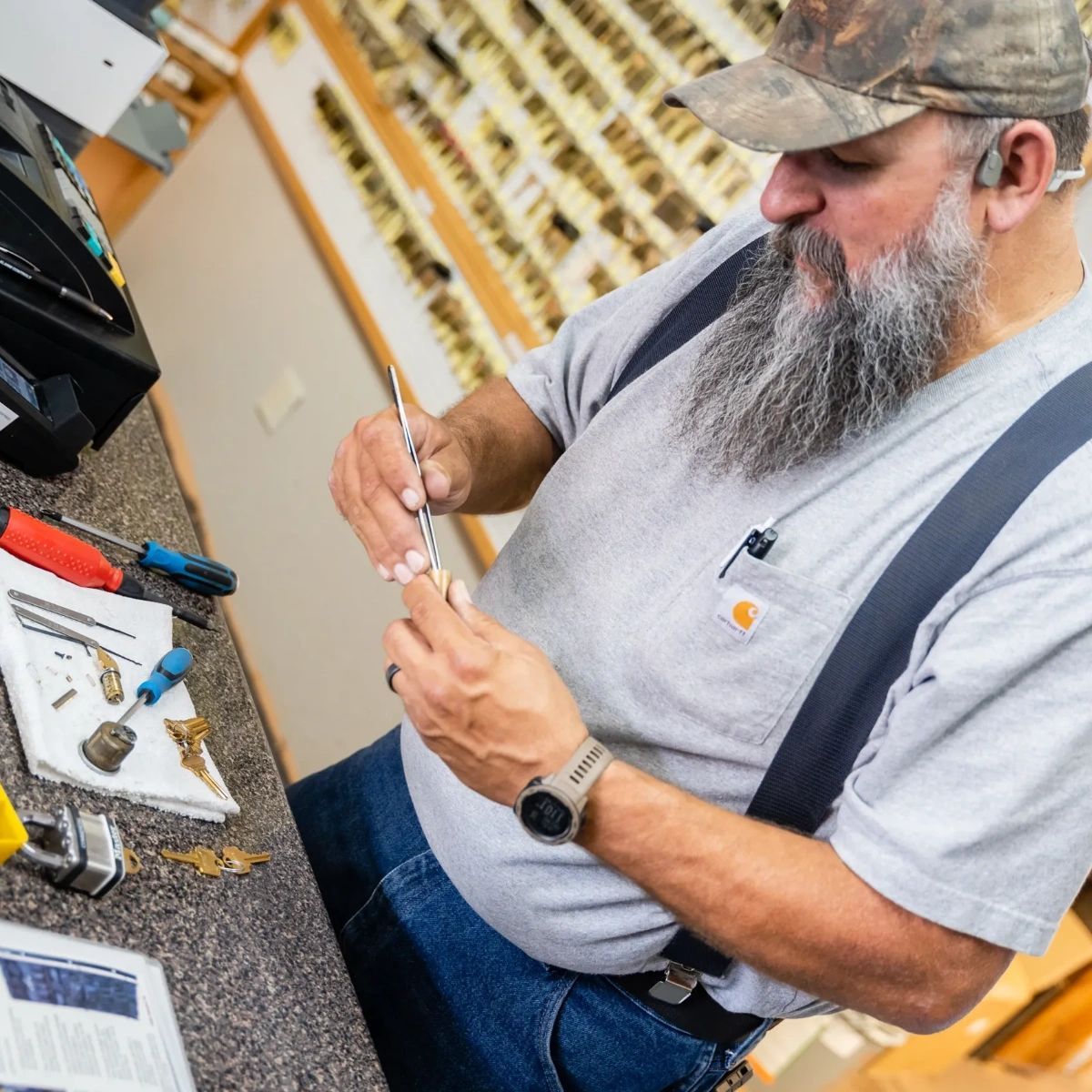 A bearded locksmith in a cap and gray shirt works on a lock component at a counter in a shop filled with keys.