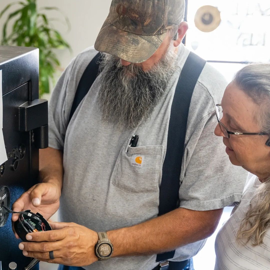 A person with a beard and camo hat demonstrates a small electrical component to another person in a workspace.