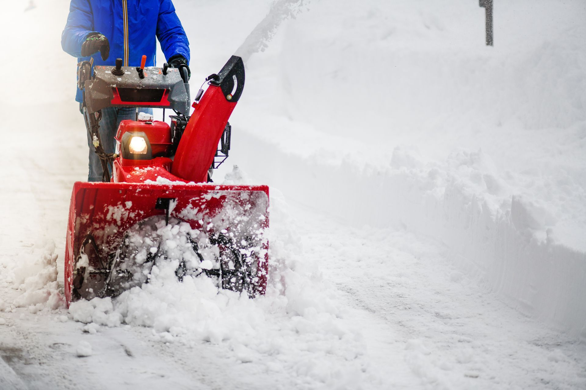 A person in a blue jacket uses a red snowblower to clear a path through deep snow.