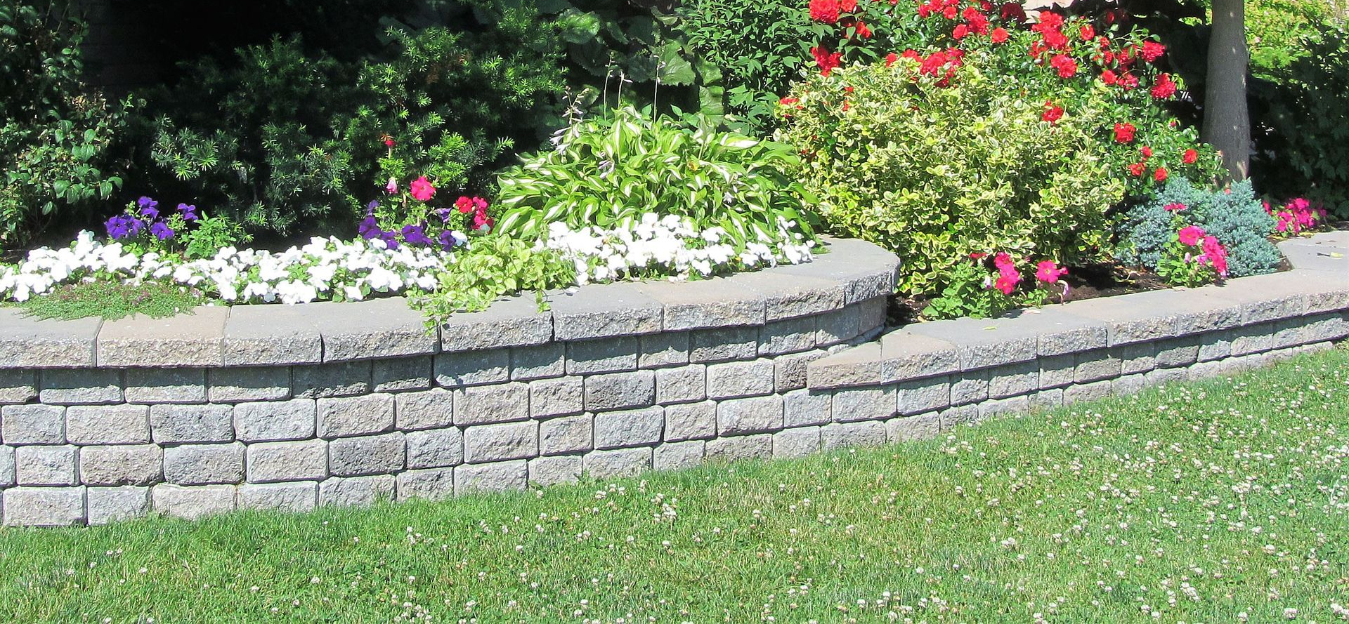 Curved stone garden bed with colorful flowers and green shrubs beside a lawn.
