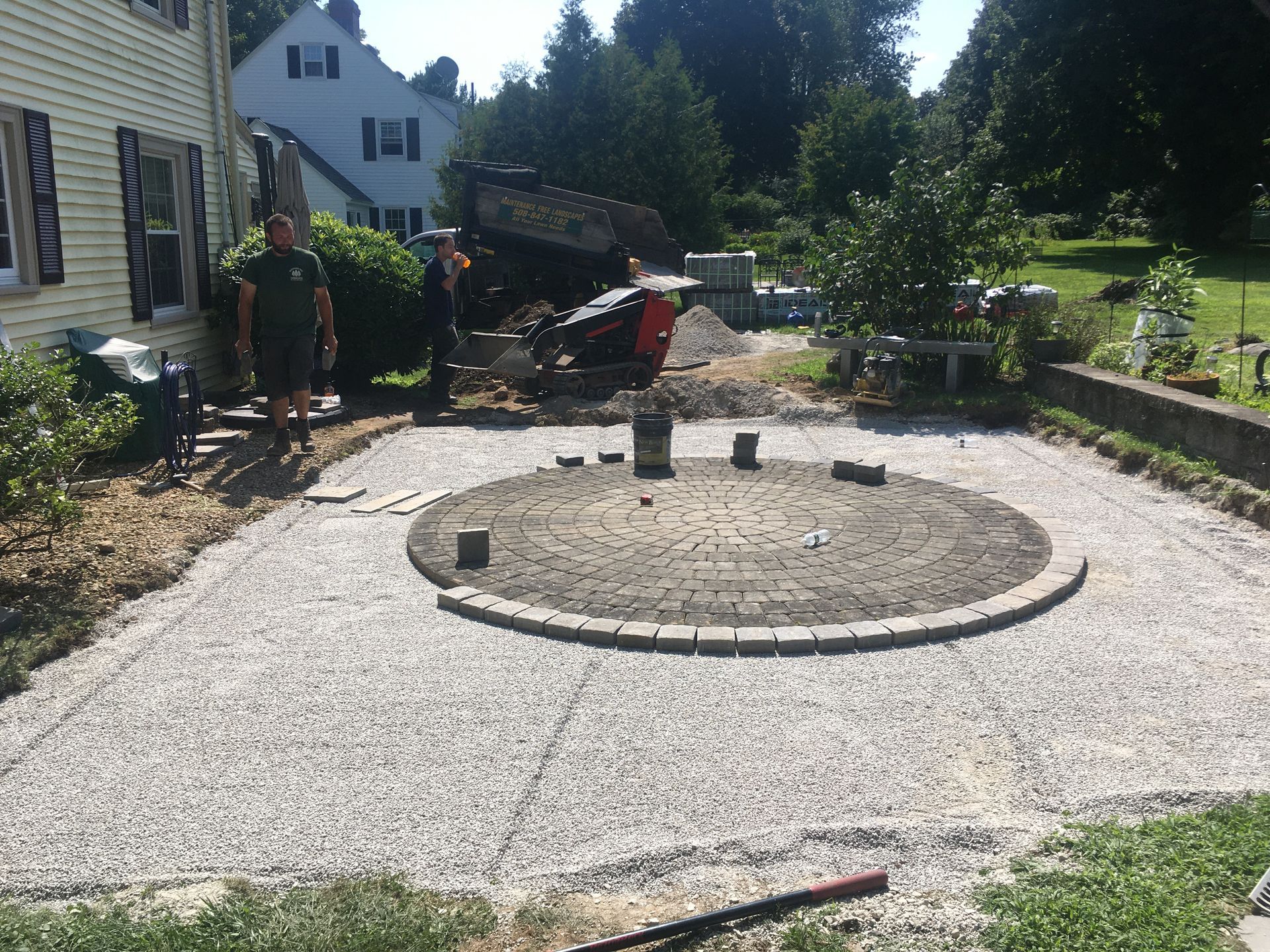 Construction workers stand near a partially completed circular stone patio set in a gravel-filled yard next to a house.