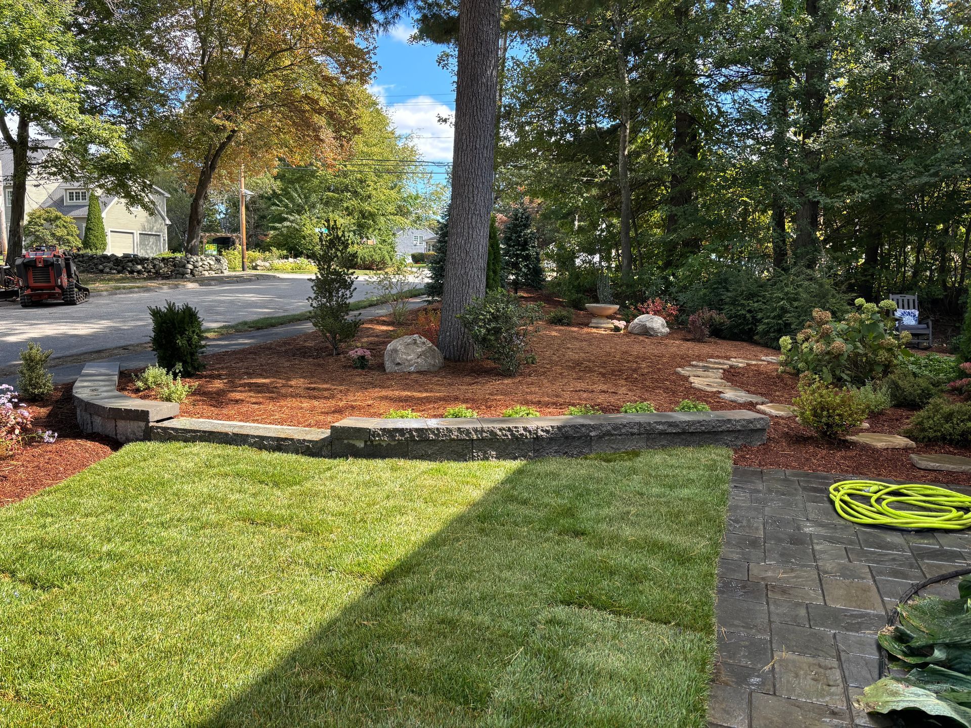A landscaped front yard featuring a stone retaining wall, fresh mulch, newly laid sod, and a large tree by the road.
