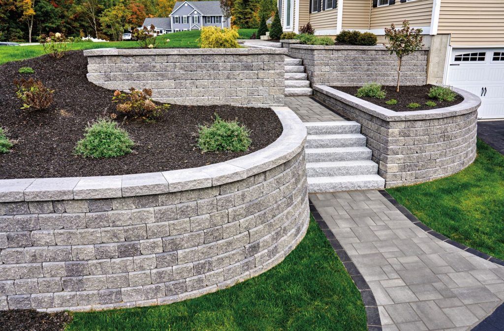 A tiered backyard landscape features stacked stone retaining walls, green plants, and a mowed lawn against a wooden fence.