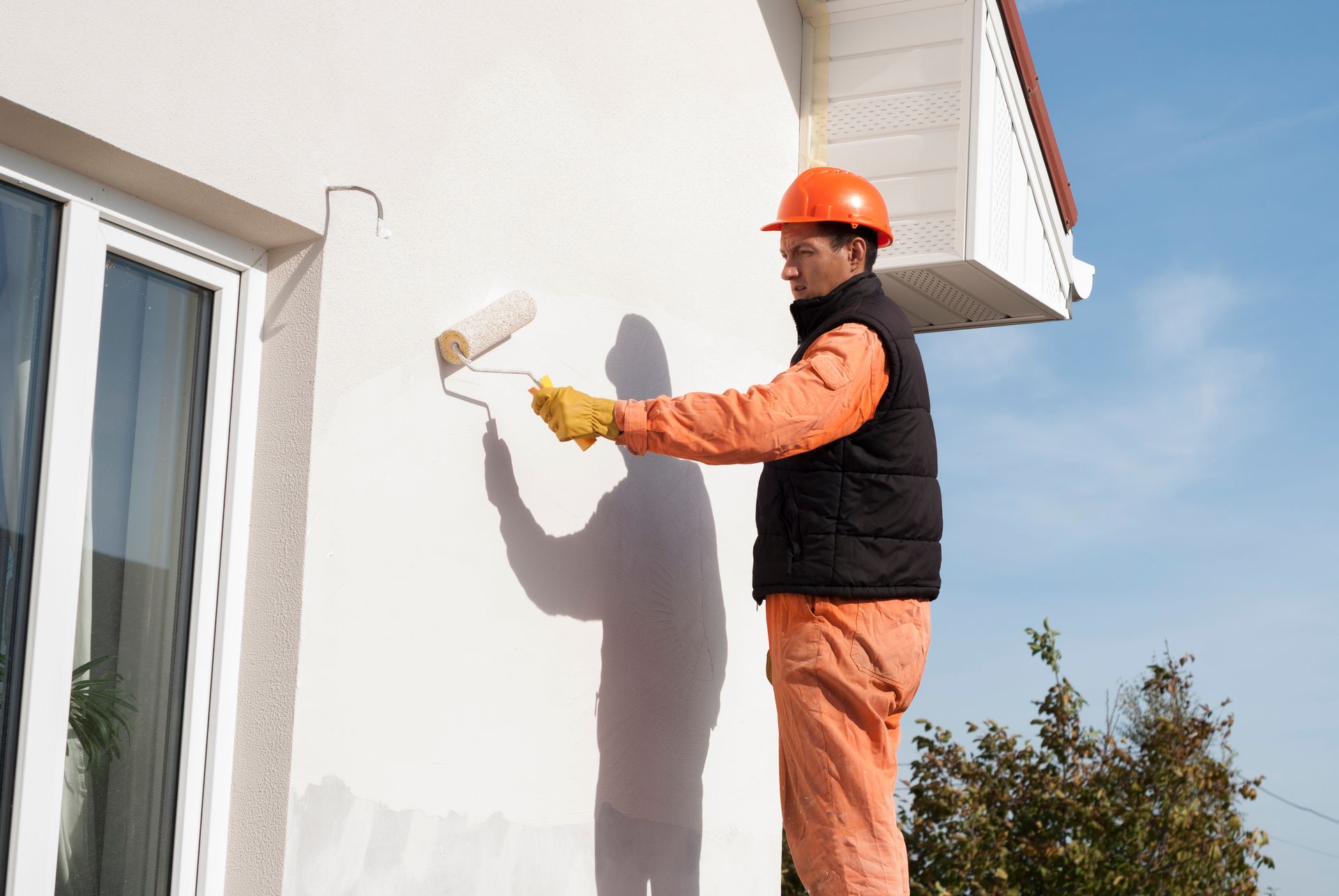 A house painter wearing work clothes and a helmet paints the outside of a home.