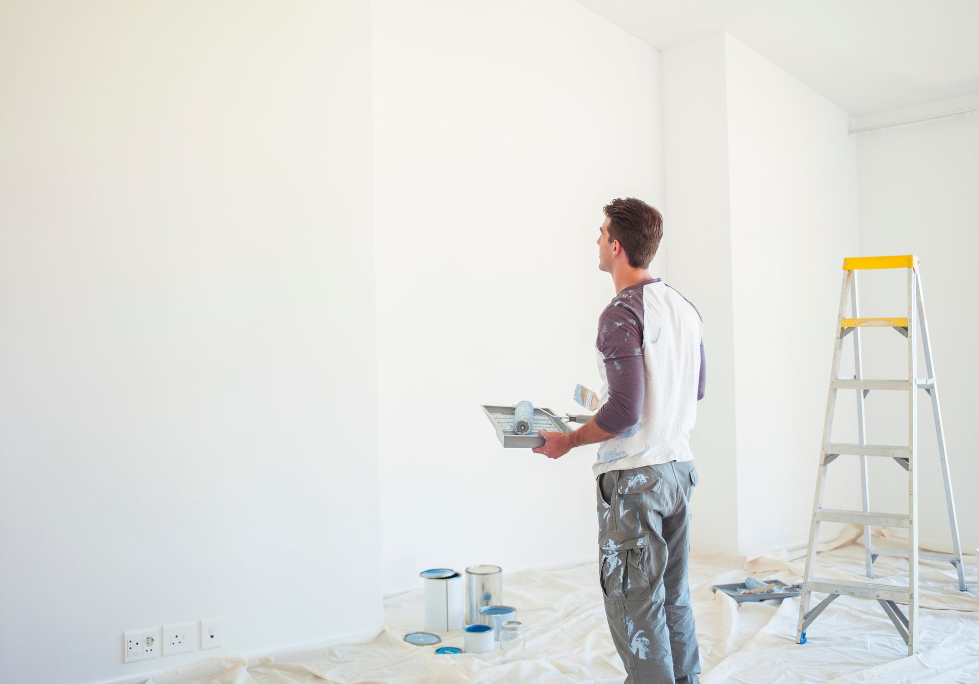 Man prepares to paint a room, holding a tray, with house painters' tools like a ladder nearby