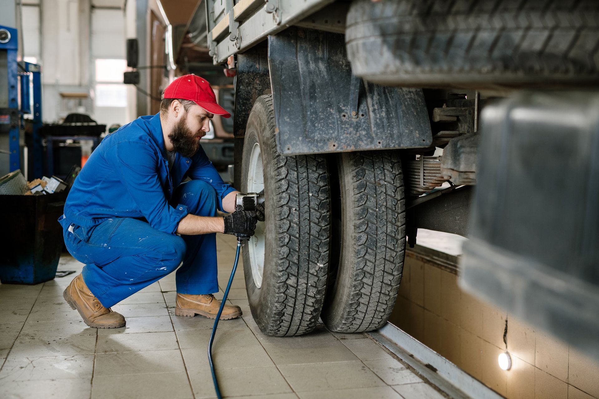 Mechanic in blue overalls and red cap inflating truck tires in a garage.