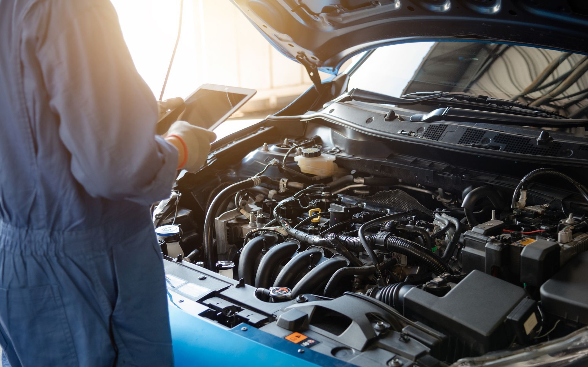 Mechanic in blue overalls examines car engine with tablet in bright garage.