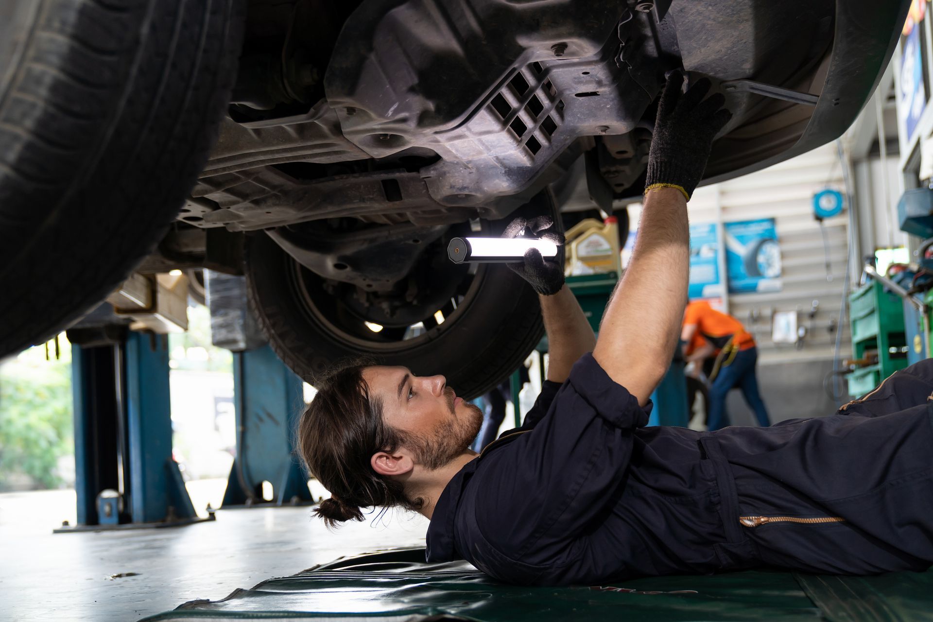 Mechanic in blue overalls working under a car, holding a light, in a garage.