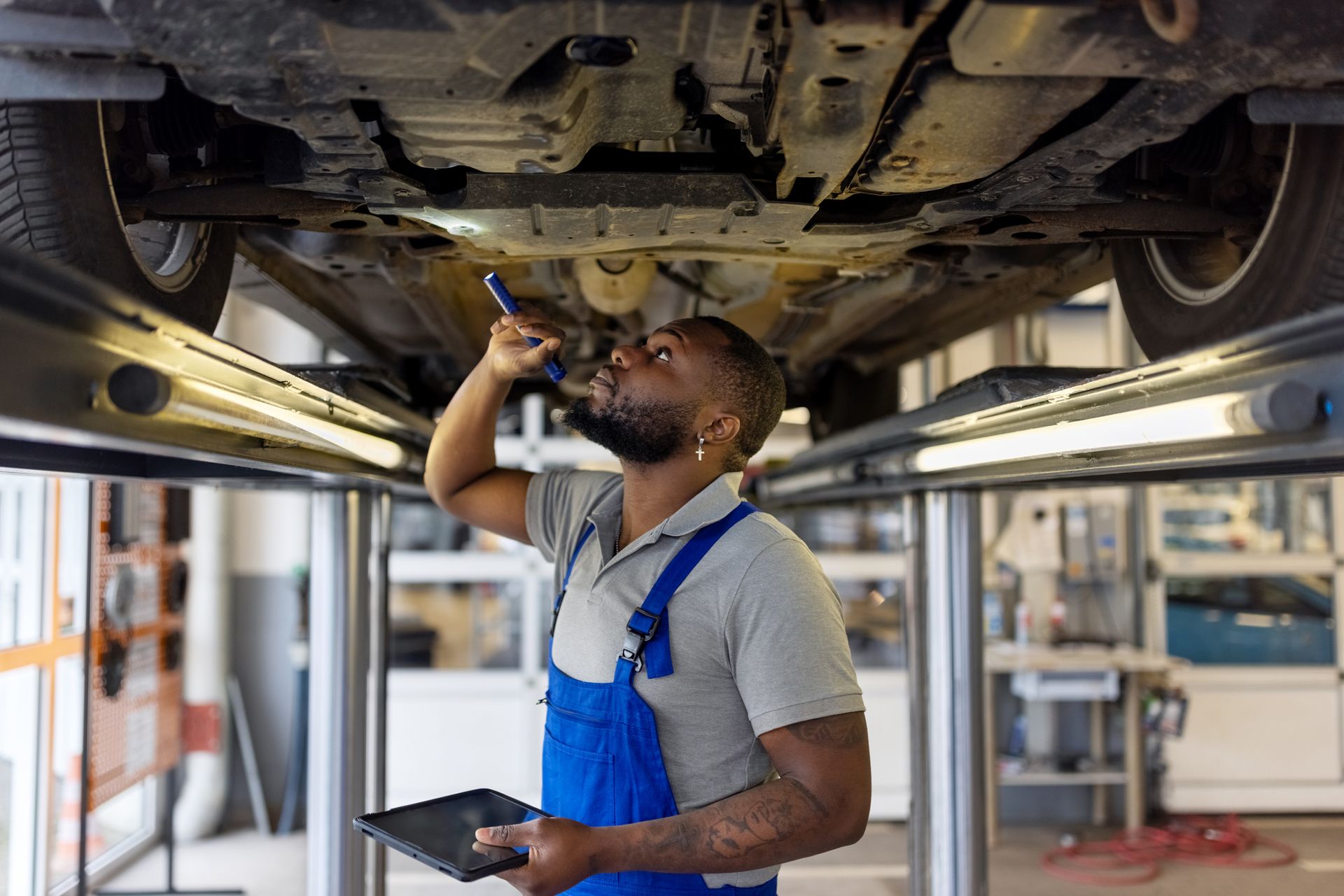 Mechanic examines car's undercarriage with flashlight, holding tablet. Garage setting, man in blue overalls.