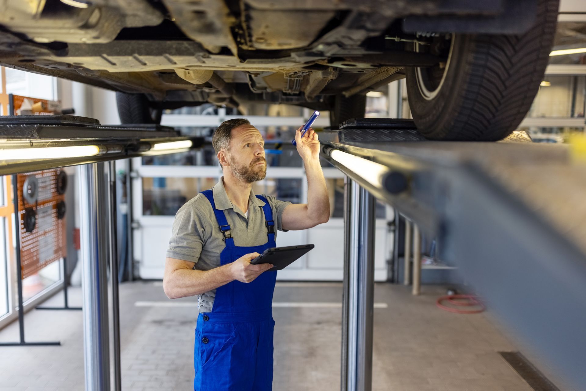 Mechanic in blue overalls inspecting car undercarriage, holding a tablet, in a garage.