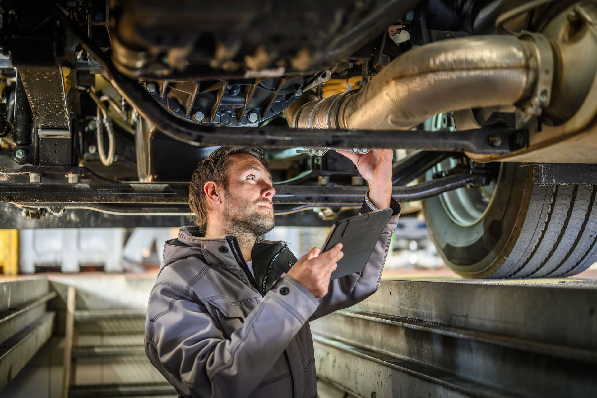 Mechanic inspecting underside of a truck, holding a tablet, in a garage.