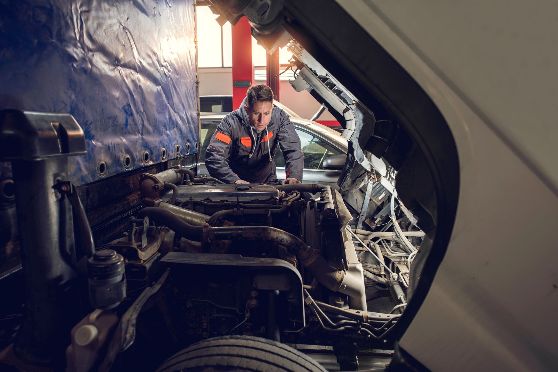 Mechanic repairs a truck engine in a garage. He wears an orange uniform and focuses intently.