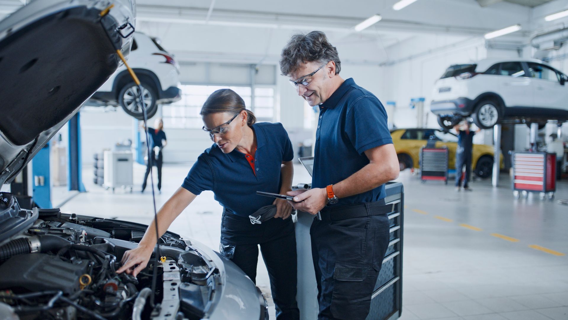 Two mechanics in blue uniforms examine a car engine in a garage.