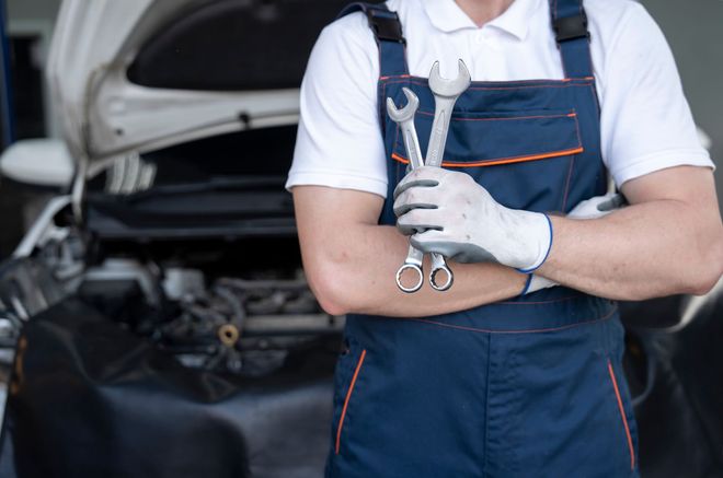 Mechanic in blue overalls holding wrenches, standing in front of a car with the hood open.