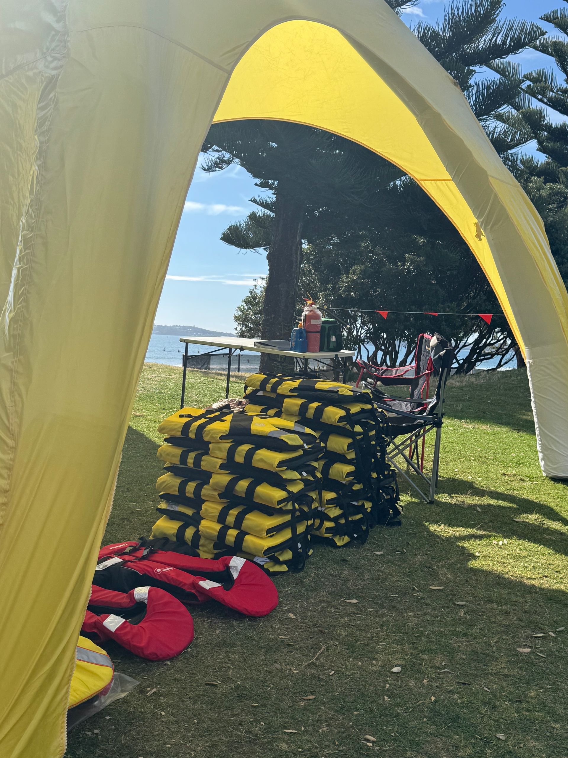 Yellow tent on a grassy area, stacked life vests and a table near the water.
