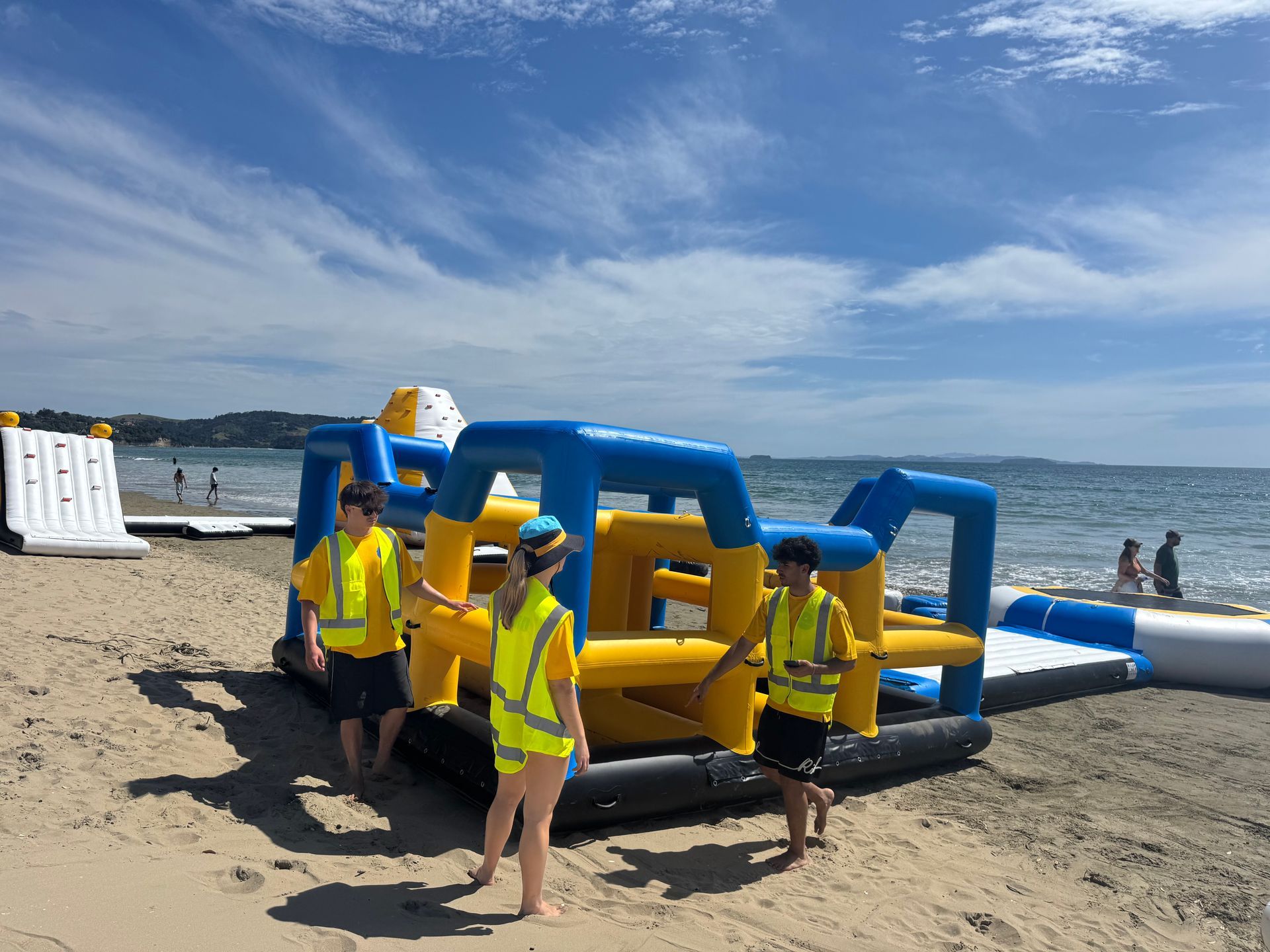 Three people in yellow vests on a sandy beach near an inflatable water obstacle course on a sunny day.
