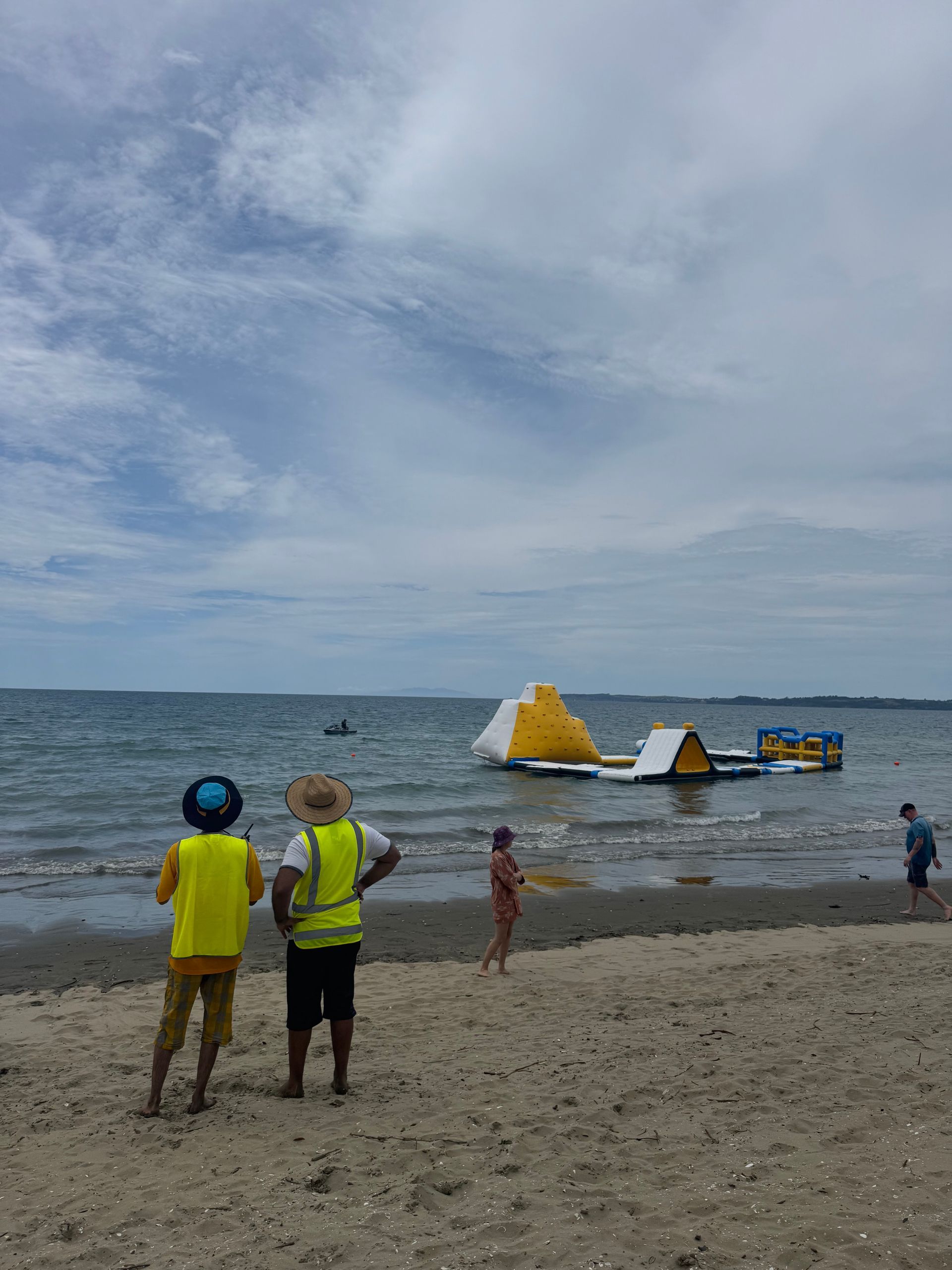 Two people on a beach watch a floating water park in the ocean under a cloudy sky.