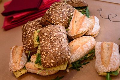 Stapel sandwiches op tafel, sommige doormidden gesneden. Brood met zaden, groene vulling. Rode servetten links.