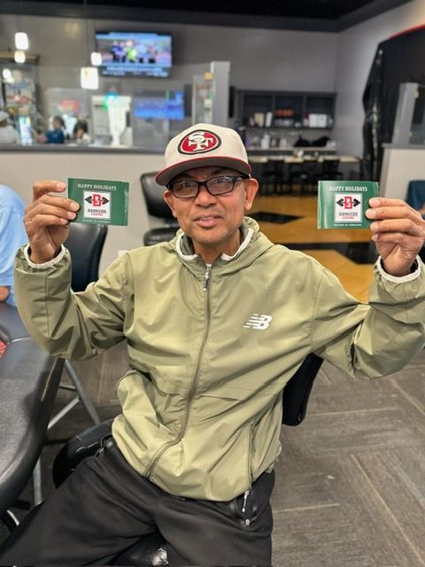 Man holding two green gift cards, smiling, wearing a jacket and hat in a lounge setting.