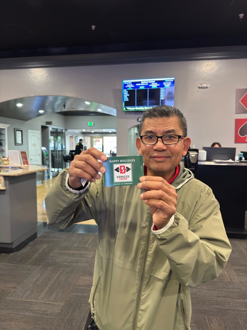 Man holding a green card, smiling. Inside a building with a TV, desk, and gray carpet.
