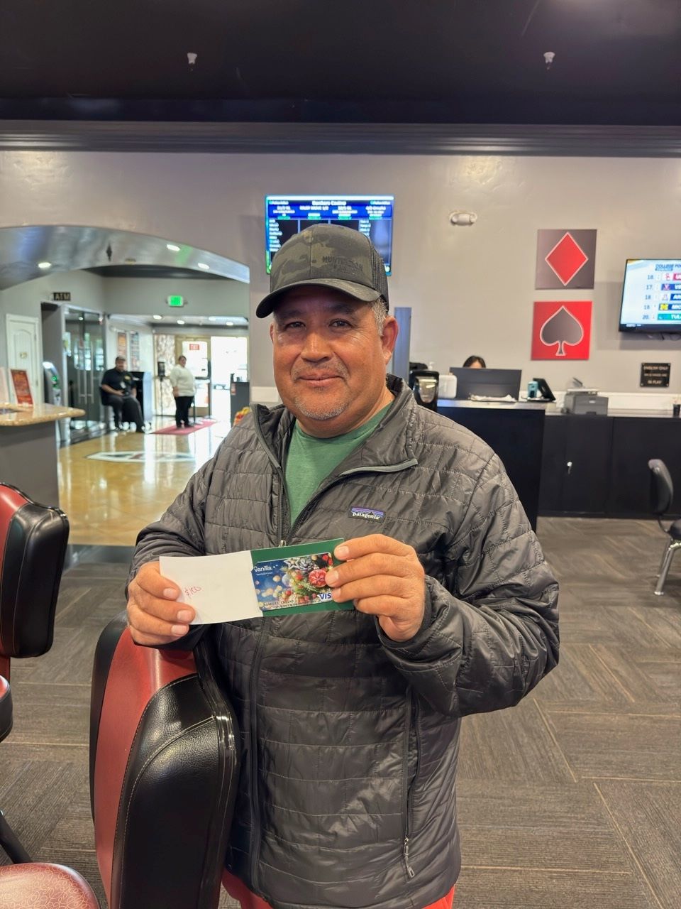 Man holding a winning ticket, smiling, in a casino setting, wearing a black cap and jacket.