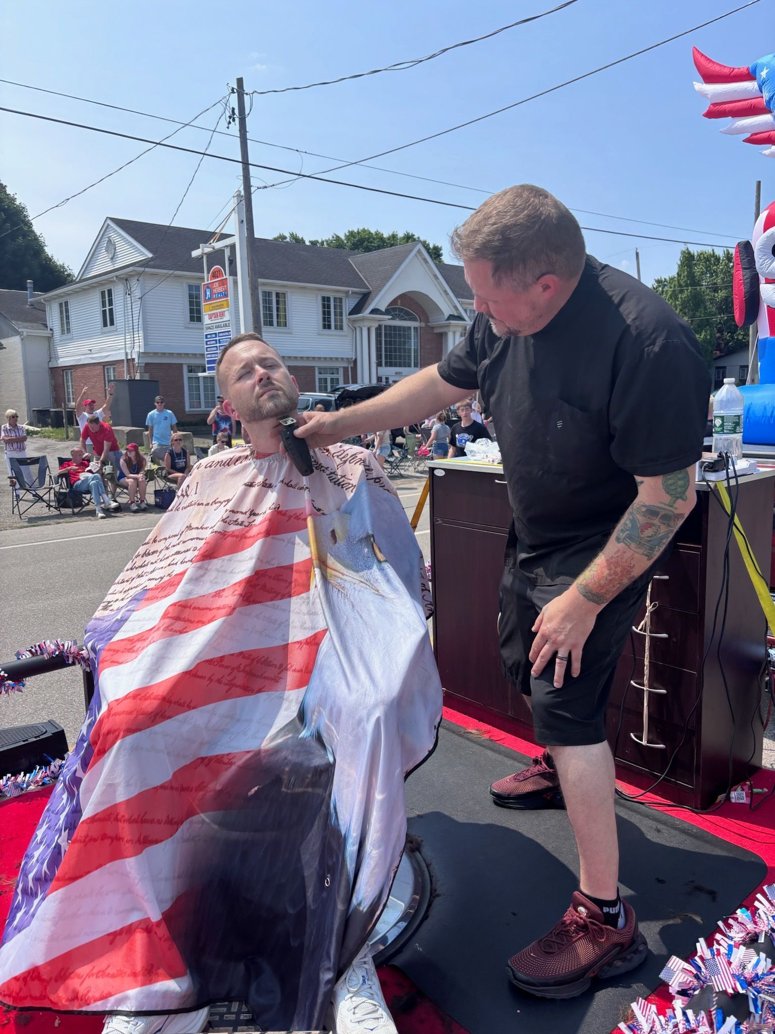 Barber shaving a seated person draped in an American flag cape at an outdoor community event