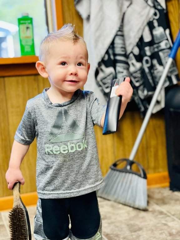 Toddler holding a small brush and bottle in a room, wearing a gray Reebok shirt and black pants
