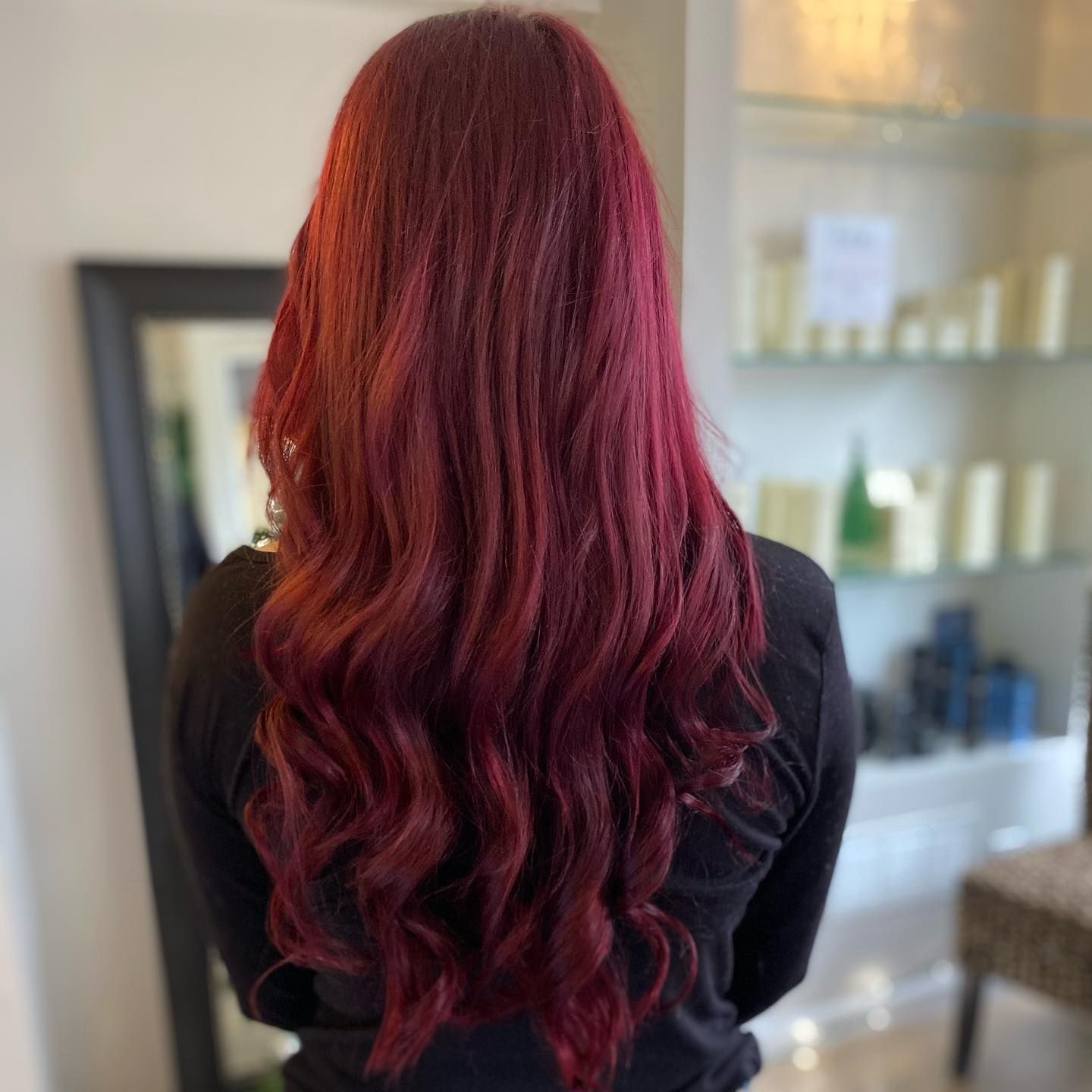 Woman with long, wavy, crimson red hair. She is wearing a black top, indoors with a mirror and shelves.