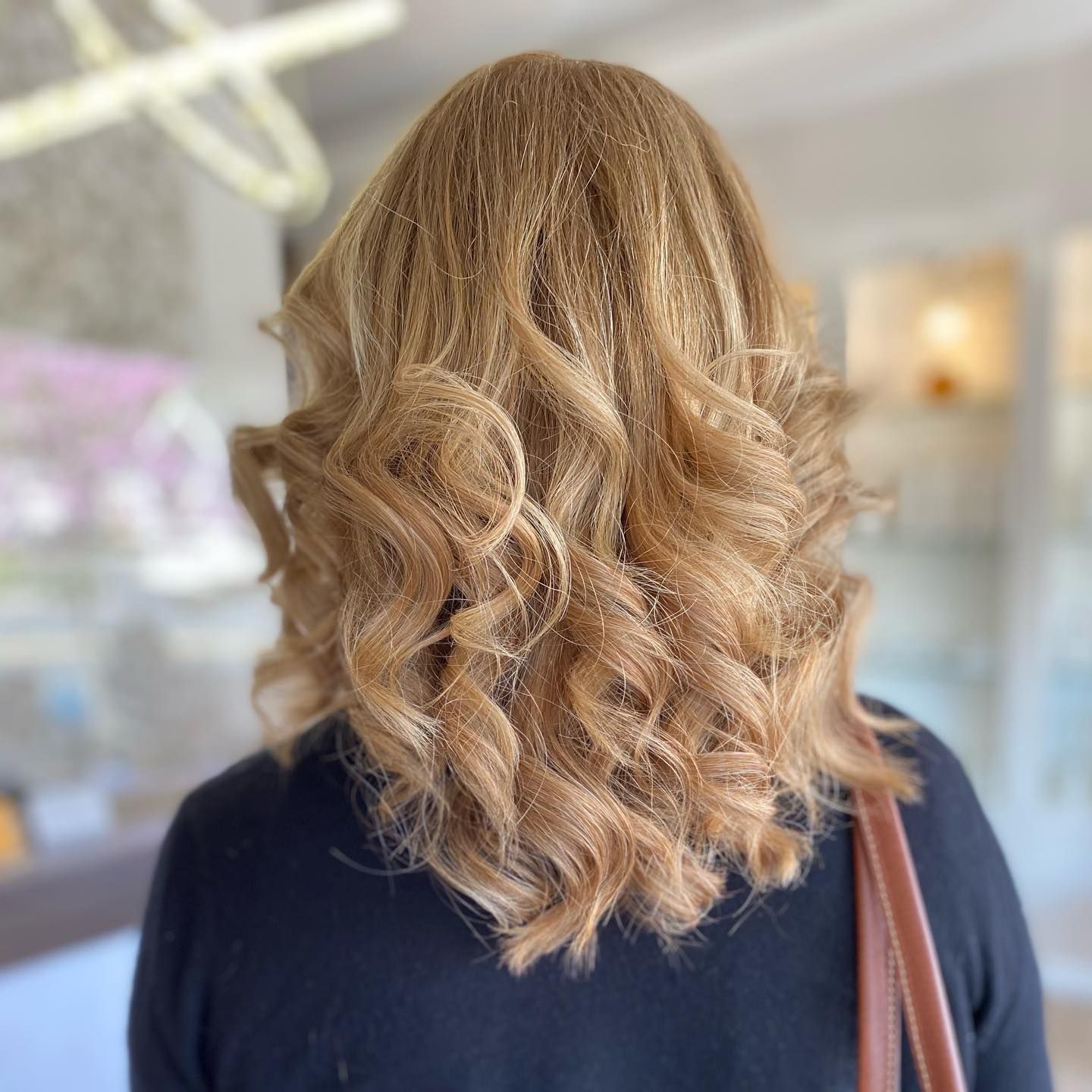 Woman with wavy, blonde hair, viewed from the back, wearing a black top, and carrying a tan purse.