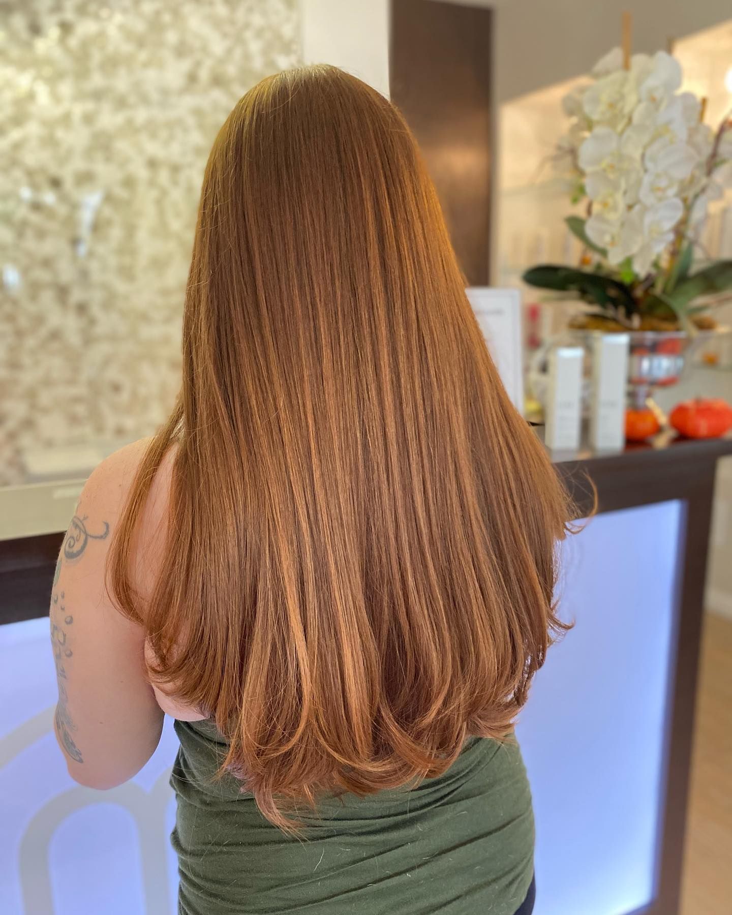 Long, copper-colored hair in a crimped style. Person stands near a counter with a flower arrangement.